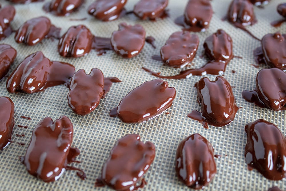 chocolate covered pecans lined up with wet chocolate on a silicone baking mat
