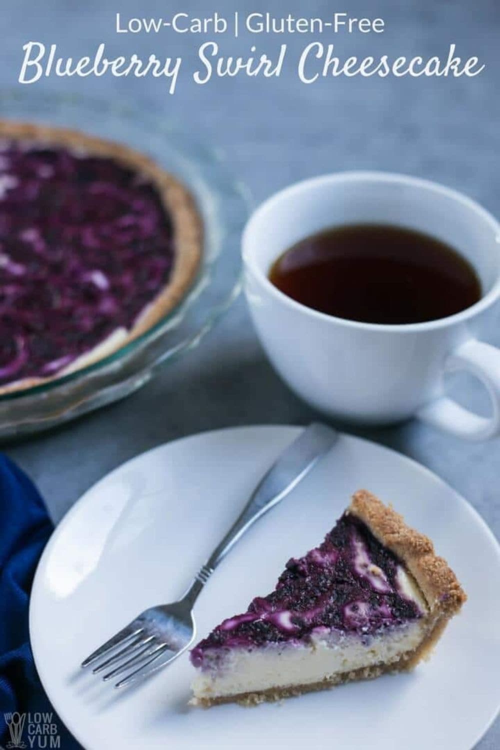 slice of blueberry swirl keto cheesecake on a white dessert dish with a fork next to it and a cup of keto coffee behind it and the blueberry swirl keto cheesecake pie dish in the background