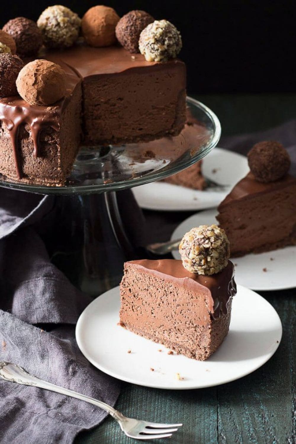 one slice of chocolate truffle keto cheesecake on a white plate with a small dessert fork next to it with a large chocolate truffle keto cheesecake sitting on a elevated glass platter behind it with a missing piece of chocolate truffle keto cheesecake