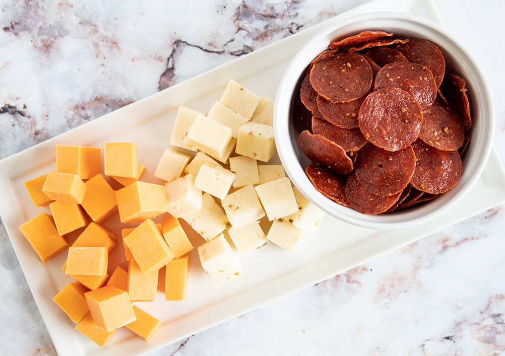 keto pepperoni in a white bowl next to two piles of cubed cheese