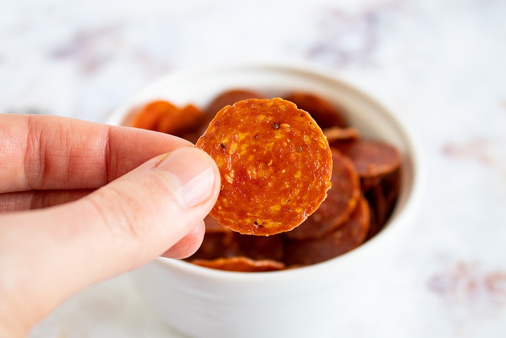 fingers holding up keto pepperoni chips in front of a bowl