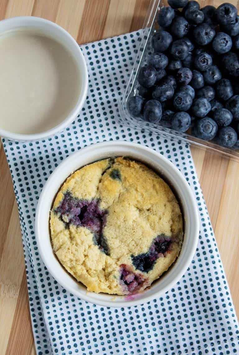 overhead view of keto blueberry mug cake on a blue and white cloth next to blueberries and icing