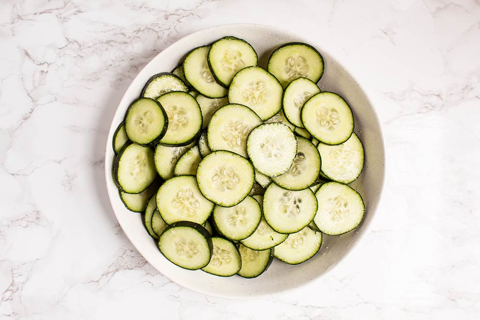 overhead view of large bowl containing sliced cucumbers seasoned with salt