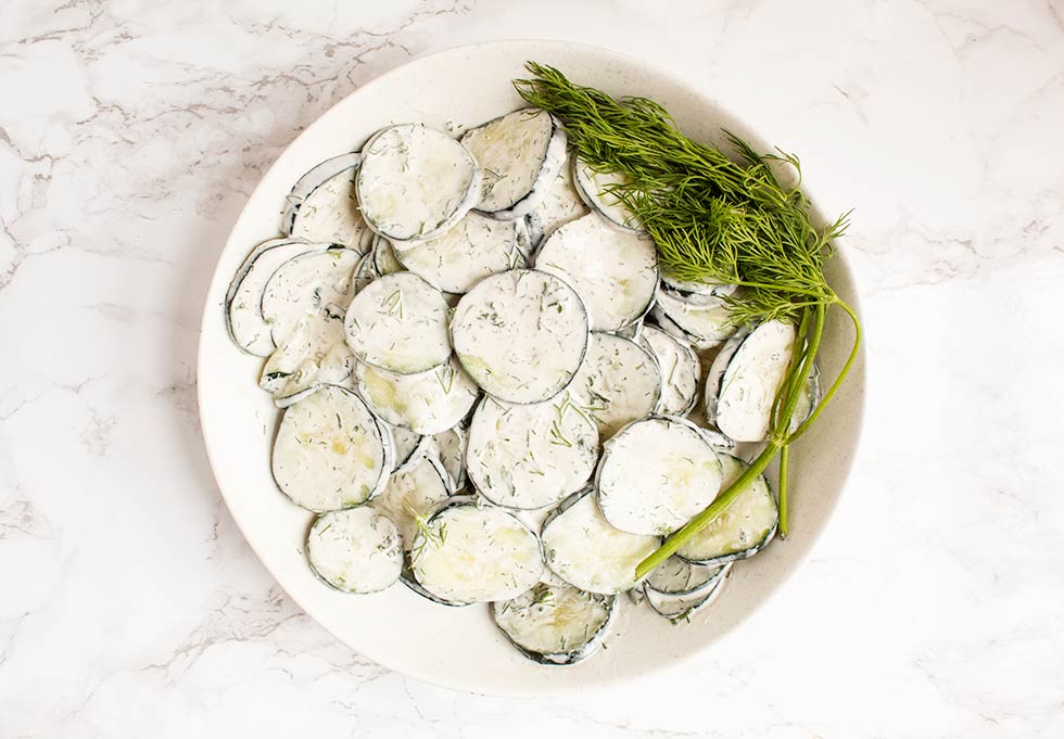 overhead view of large off-white bowl containing cucumbers with dill on the side