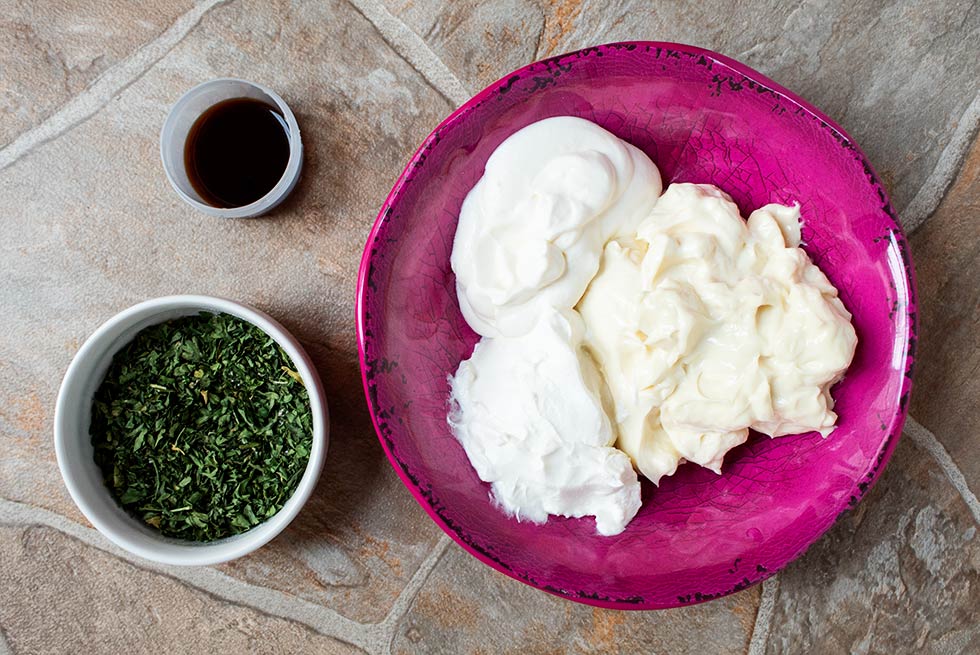 overhead view of pink bowl with keto ranch dressing recipe ingredients next to a bowl of herbs