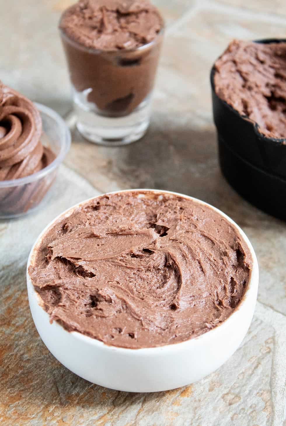 closeup of keto cream cheese chocolate frosting in a white bowl with keto frosting in various containers behind it