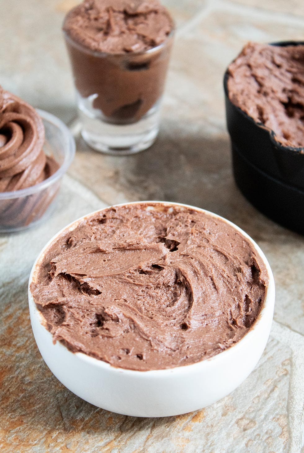 closeup of keto chocolate frosting in a white bowl with keto frosting in various containers behind it