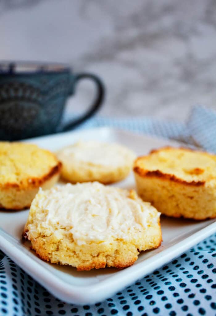 sideview of four cornbread muffings and one has butter all on a white serving plate on a blue tablecloth and a dark blue mug in background