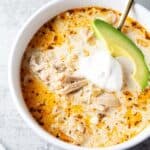 overhead view of bowl of keto white chicken chili in white serving bowl and gold serving spoon and garnished with slice of avocado and sour cream
