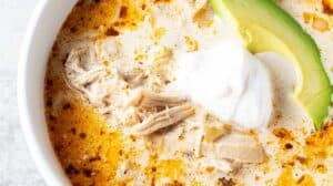 overhead view of bowl of keto white chicken chili in white serving bowl and gold serving spoon and garnished with slice of avocado and sour cream