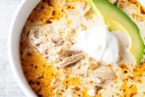 overhead view of bowl of keto white chicken chili in white serving bowl and gold serving spoon and garnished with slice of avocado and sour cream