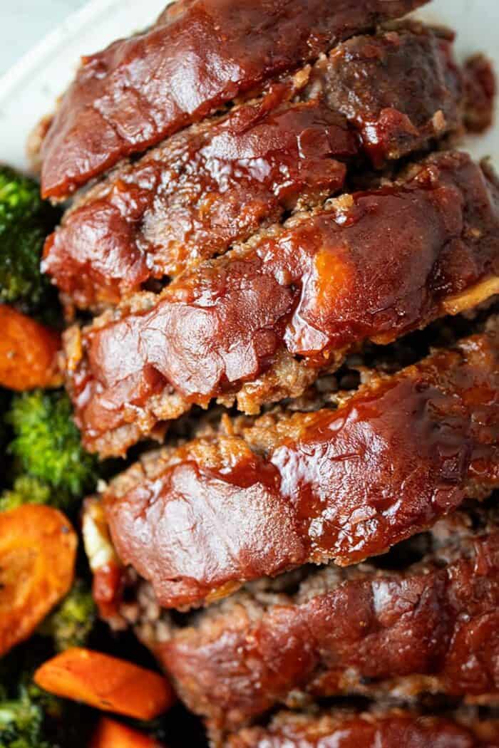 overhead view of sliced keto meatloaf with ketchup glaze on a white serving dish and carrots and broccoli on the side